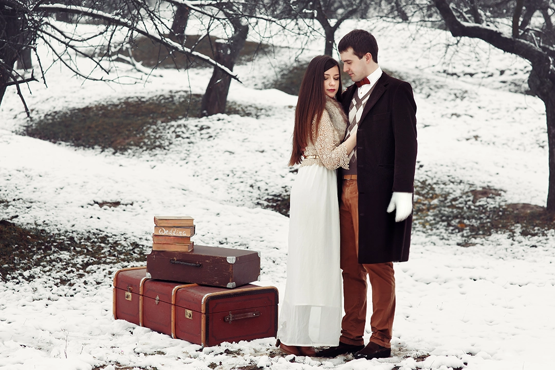 Couple de mariés posant dans la neige avec des valises vintage lors d'un mariage d'hiver.